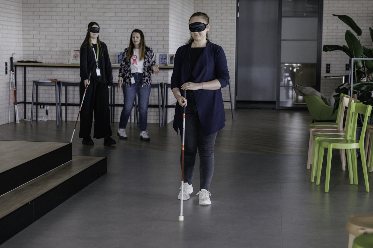 Two blindfolded women move around the office space with white canes. A third person, a mentor from the Foundation, stands behind and instructs the participants.